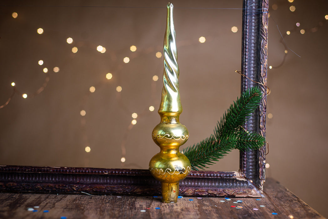A golden Christmas ornament with a pointed top and a sprig of greenery is placed on a wooden surface, with a blurred background of twinkling lights.