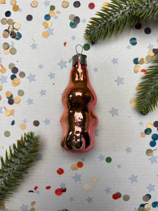 A shiny, metallic ornament in the shape of a bottle is surrounded by confetti and pine branches on a white background.