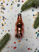 A shiny, metallic ornament in the shape of a bottle is surrounded by confetti and pine branches on a white background.