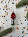 A red ornament with white polka dots is placed on a white background, surrounded by colorful confetti and green pine branches.