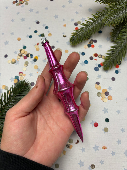 A hand holding a pink ornament with a pointed end, surrounded by confetti and pine branches.
