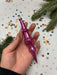 A hand holding a pink ornament with a pointed end, surrounded by confetti and pine branches.