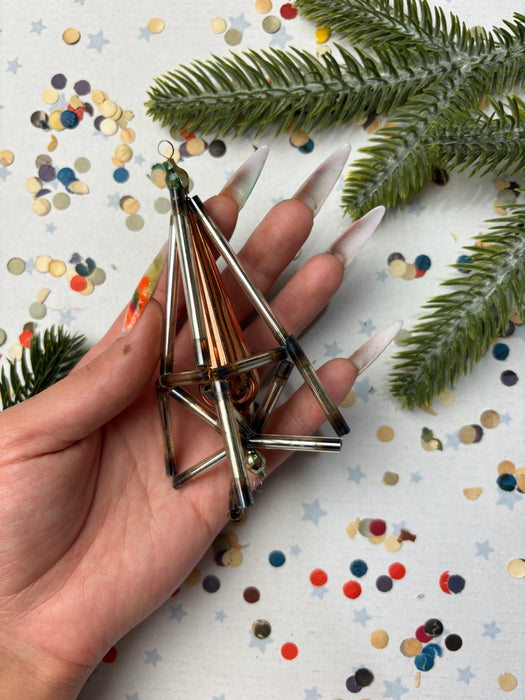 A hand holding a metallic ornament with a triangular shape, surrounded by confetti and a pine tree branch.
