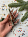 A hand holding a metallic ornament with a triangular shape, surrounded by confetti and a pine tree branch.
