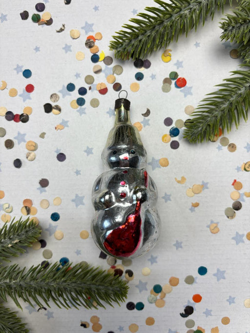 A silver snowman ornament with a red scarf is surrounded by colorful confetti and pine branches on a white background.