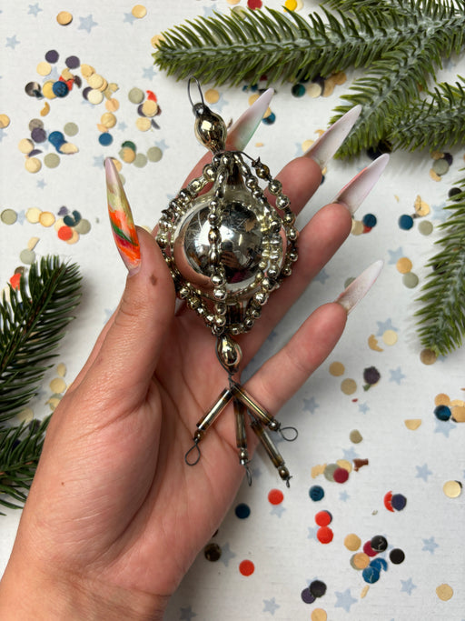 A hand holding a decorative ornament with a metallic, ornate design, surrounded by confetti and pine branches.