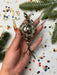 A hand holding a decorative ornament with a metallic, ornate design, surrounded by confetti and pine branches.