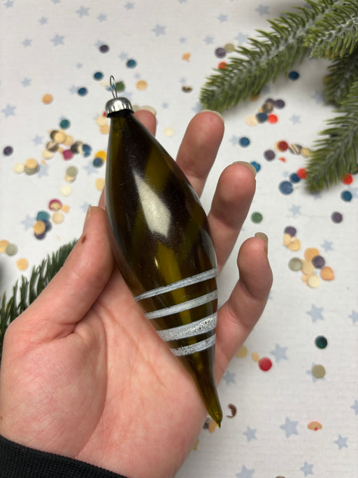 A hand holding a green glass ornament with white stripes, surrounded by colorful confetti and a pine tree branch.