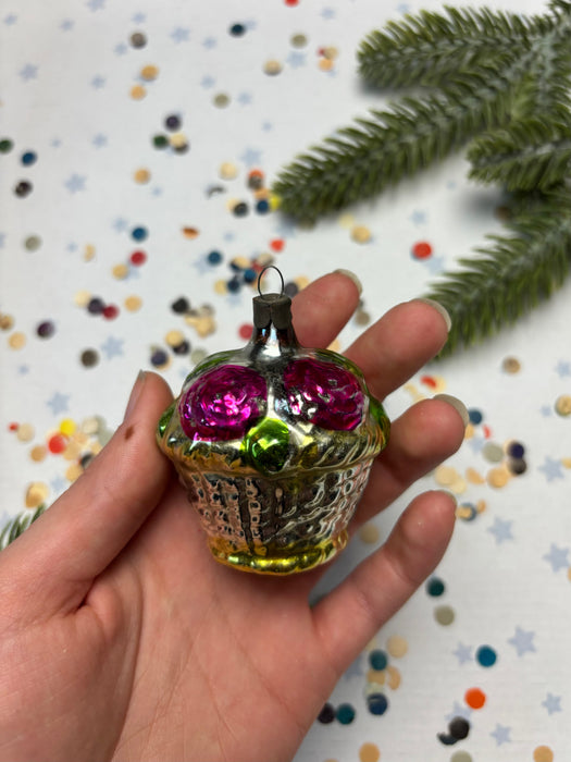 A hand holding a small, colorful ornament with a cupcake-like design, surrounded by confetti and a pine branch.