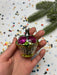 A hand holding a small, colorful ornament with a cupcake-like design, surrounded by confetti and a pine branch.
