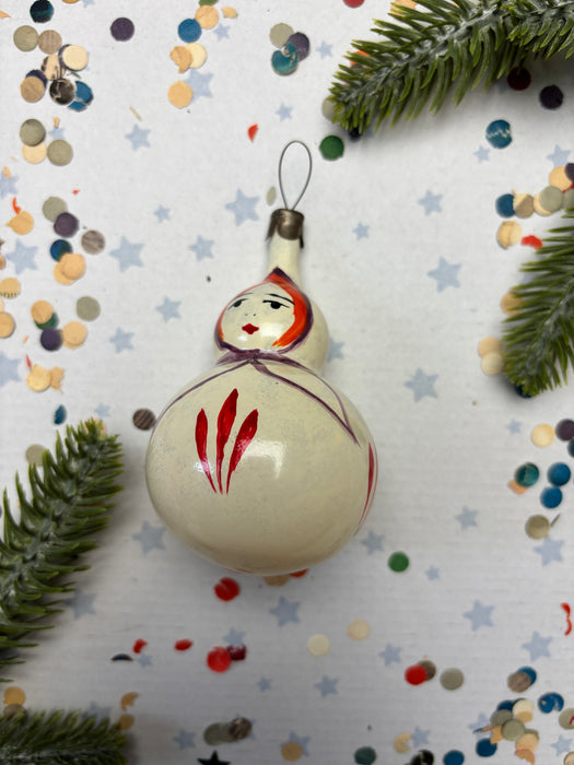 A white ornament with red and purple accents is placed on a white background, surrounded by scattered confetti and pine branches.