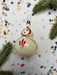 A white ornament with red and purple accents is placed on a white background, surrounded by scattered confetti and pine branches.