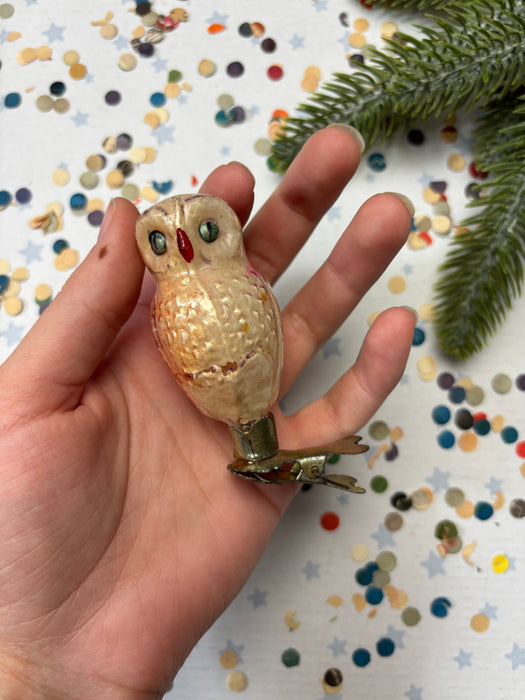 A hand holding a small, beige-colored ornament resembling an owl, with a metallic base and a red dot on the eye. The background is a white sheet with colorful confetti scattered across it.
