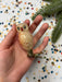 A hand holding a small, beige-colored ornament resembling an owl, with a metallic base and a red dot on the eye. The background is a white sheet with colorful confetti scattered across it.