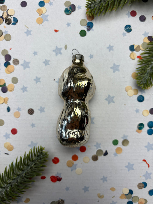 A shiny silver ornament is placed on a white background, surrounded by colorful confetti and a sprig of greenery.
