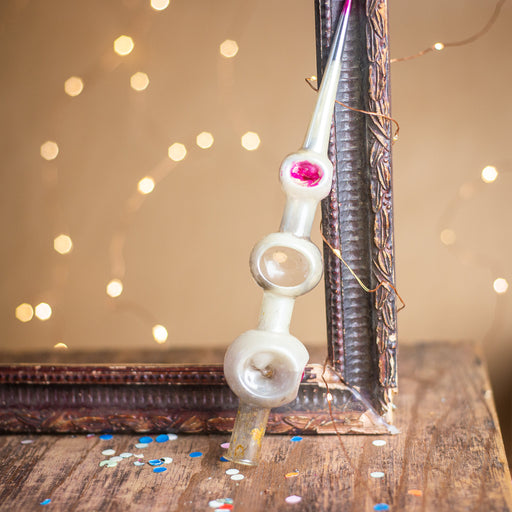 a decorative glass ornament with a pink center, leaning against a wooden frame with a decorative design. The ornament is placed on a wooden surface, with scattered confetti around it.