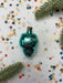 A turquoise Christmas ornament is surrounded by colorful confetti and a sprig of greenery on a white background.