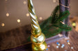 A golden Christmas ornament with a spiral design sits on a wooden surface, surrounded by a blurred background of twinkling lights.