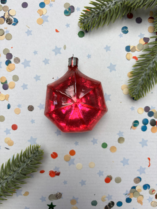 A red glass ornament with a star-shaped design sits on a white background, surrounded by scattered confetti and pine branches.