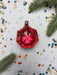 A red glass ornament with a star-shaped design sits on a white background, surrounded by scattered confetti and pine branches.