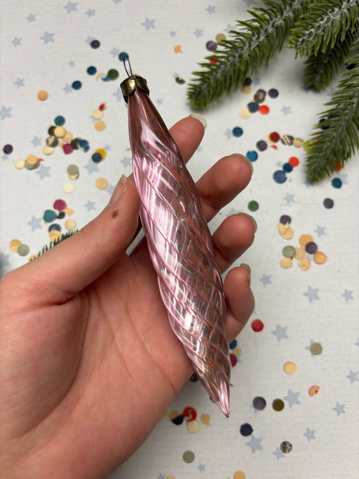 A hand holding a pink spiral ornament against a background of colorful confetti and a pine tree branch.