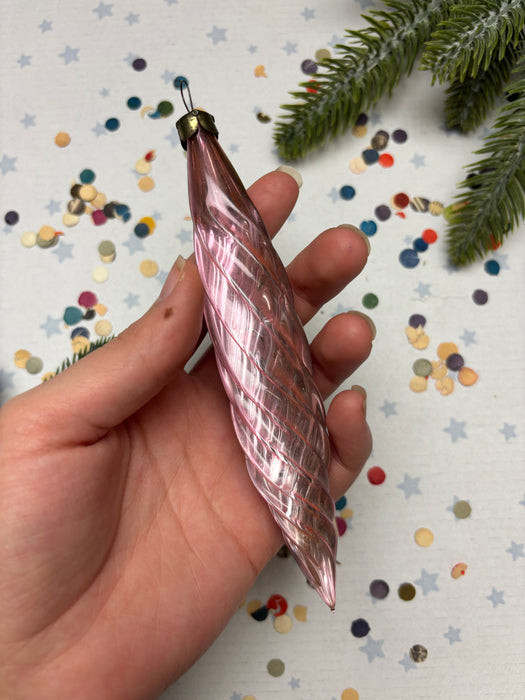 A hand holding a pink spiral ornament against a background of colorful confetti and a pine tree branch.