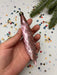A hand holding a pink spiral ornament against a background of colorful confetti and a pine tree branch.
