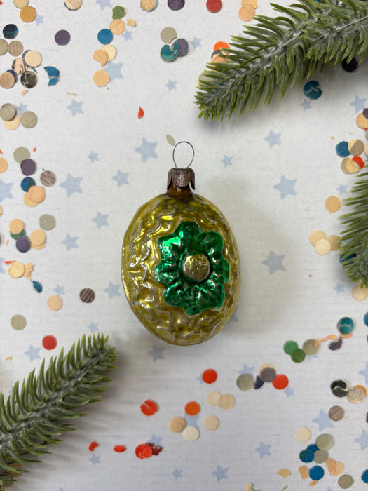 A vintage green and gold ornament with a gold flower sits on a white background, surrounded by scattered confetti and a sprig of greenery.