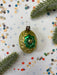 A vintage green and gold ornament with a gold flower sits on a white background, surrounded by scattered confetti and a sprig of greenery.