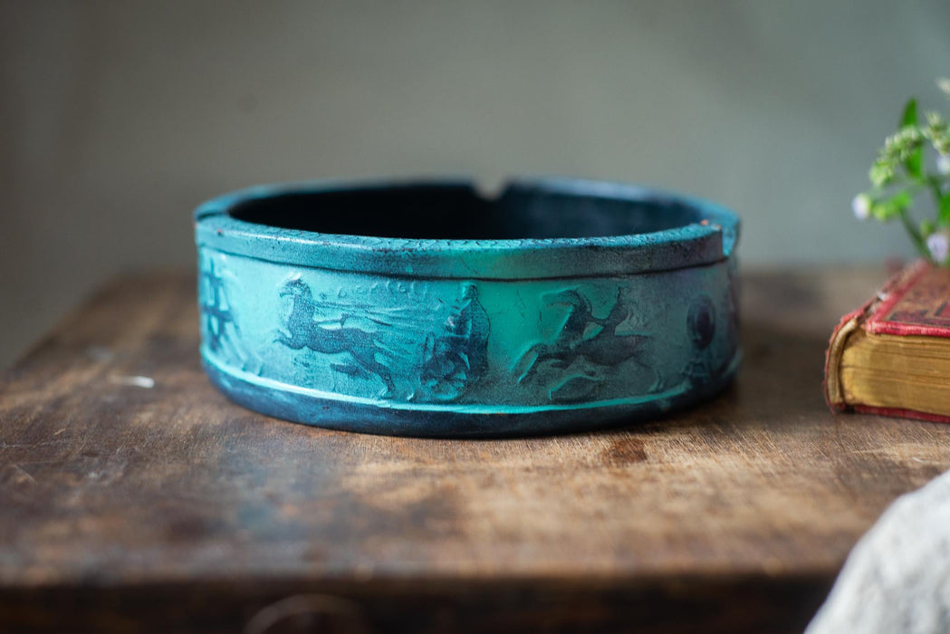 A blue ashtray with a carved design sits on a wooden surface next to a book.