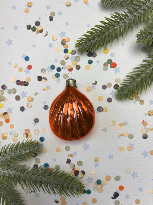 An orange glass ornament is surrounded by confetti and pine branches on a white background.