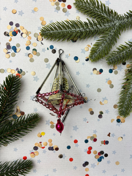 a Christmas ornament with a red ribbon and a small red ball, surrounded by pine branches, confetti, and scattered small circles on a white background.