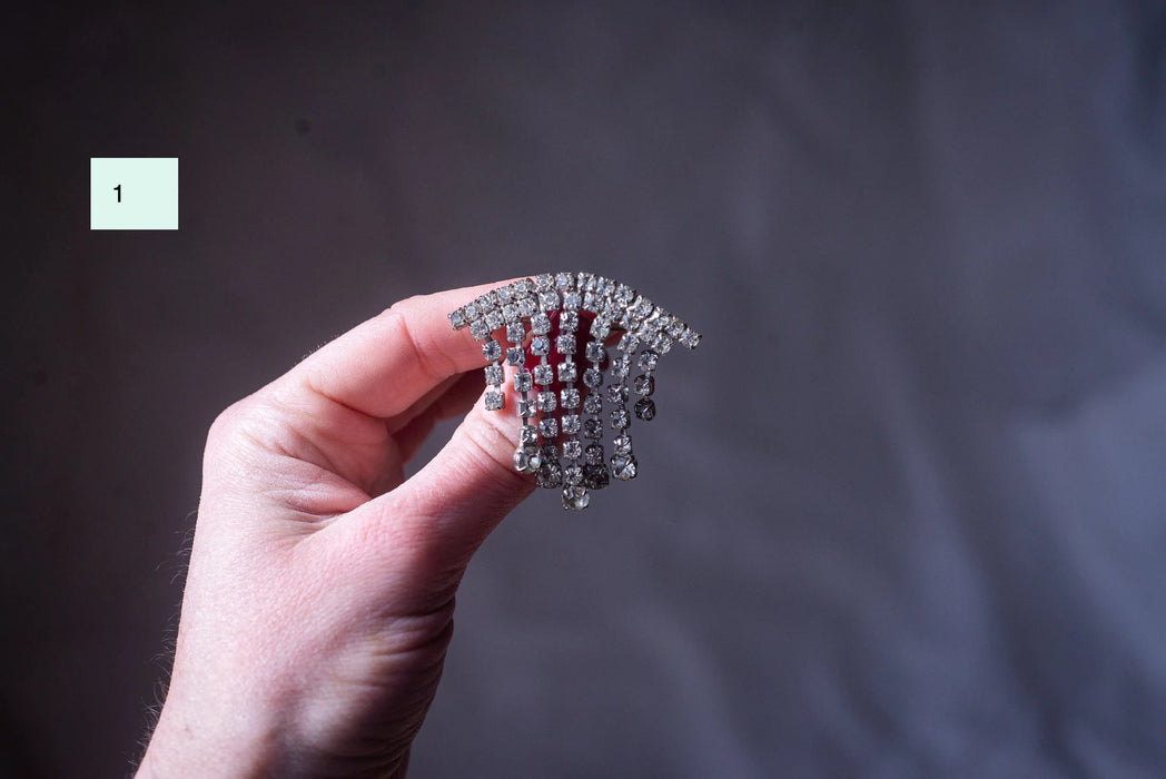 A hand holding a decorative brooch with a dangling design of small, clear stones.