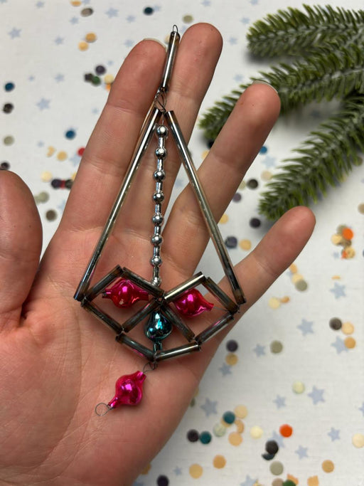A hand holding a decorative metal ornament with colorful glass ornaments, including a pink and blue glass ball, against a background of confetti and a pine tree branch.