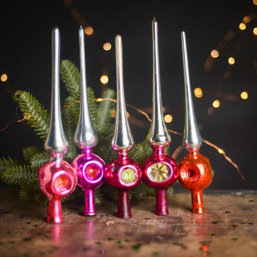 A row of colorful glass ornaments with pointed tops, arranged on a wooden surface against a dark background with twinkling lights.