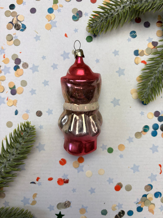 A red ornament with a bow sits on a white background, surrounded by scattered confetti and green pine branches.