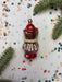 A red ornament with a bow sits on a white background, surrounded by scattered confetti and green pine branches.