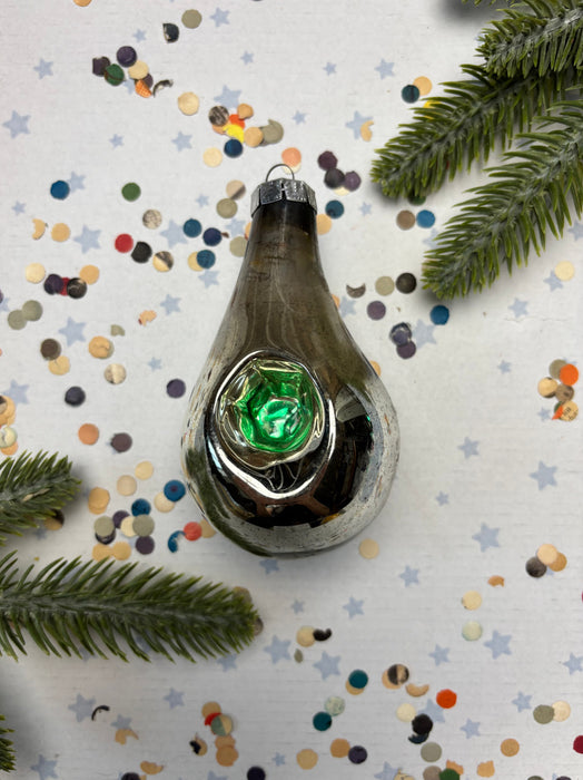 A shiny, metallic ornament with a green gemstone is surrounded by colorful confetti on a white background.