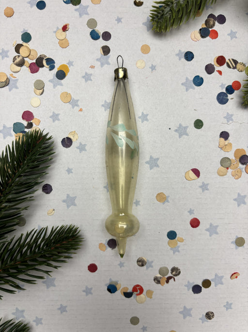 A clear glass ornament with a pointed end is placed on a white background with scattered confetti and pine branches.