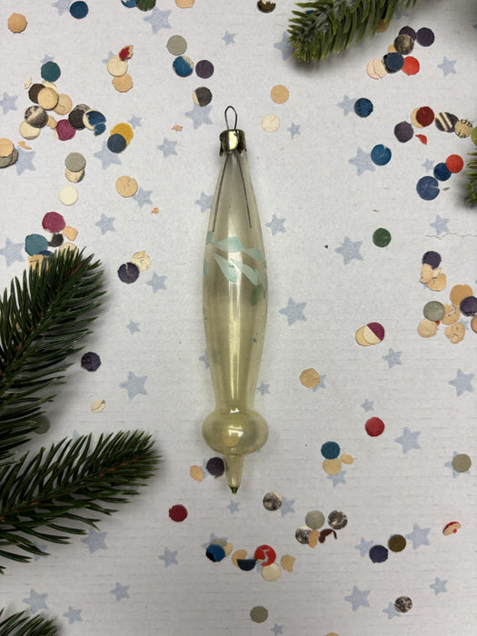 A clear glass ornament with a pointed end is placed on a white background with scattered confetti and pine branches.