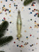 A clear glass ornament with a pointed end is placed on a white background with scattered confetti and pine branches.