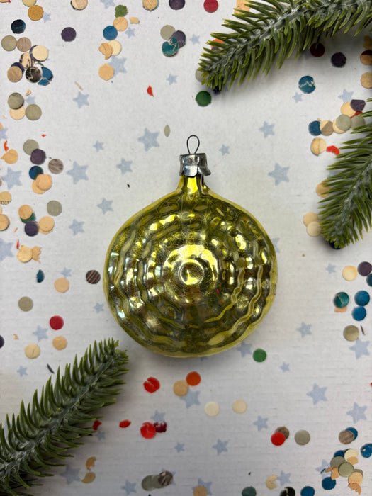 A shiny gold ornament is placed on a white background, surrounded by scattered confetti and a sprig of greenery.