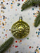 A shiny gold ornament is placed on a white background, surrounded by scattered confetti and a sprig of greenery.