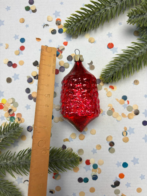 A red glass ornament is placed next to a ruler on a background of confetti and pine branches.