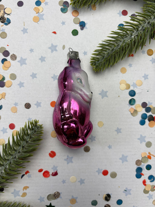 A pink glass ornament with a purple and white design sits on a white background, surrounded by colorful confetti and green pine branches.
