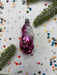 A pink glass ornament with a purple and white design sits on a white background, surrounded by colorful confetti and green pine branches.