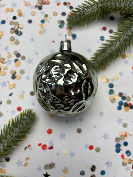 a shiny silver ornament with a reflective surface, surrounded by colorful confetti and green pine branches.