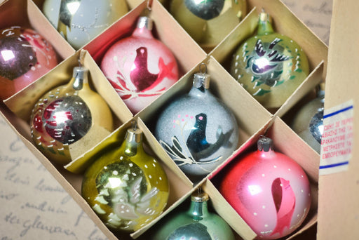 A cardboard box filled with a variety of colorful Christmas ornaments, including glass baubles, baubles with bird designs, and other festive decorations.