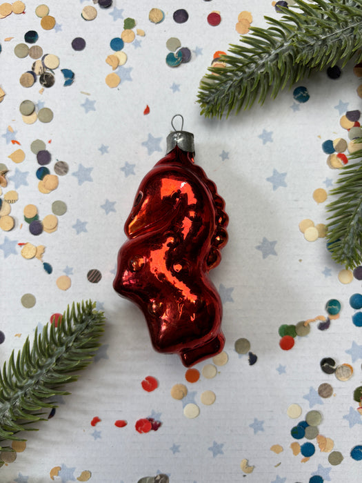 A red ornament resembling a heart shape is placed on a white background with scattered confetti and pine branches.