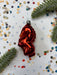 A red ornament resembling a heart shape is placed on a white background with scattered confetti and pine branches.